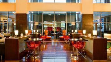 Un hall d’hôtel moderne doté d’un bar central, de sièges avec des fauteuils rouges et de grandes fenêtres offrant une vue sur la ville.