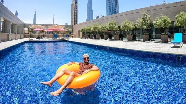 A pool area with a person on an inflatable ring, surrounded by modern buildings and trees. - King Room Near Civil Defance Downtown by E R (Dubai)