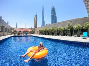 Uma área de piscina com uma pessoa em um flutuador, cercada por edifícios modernos e árvores.