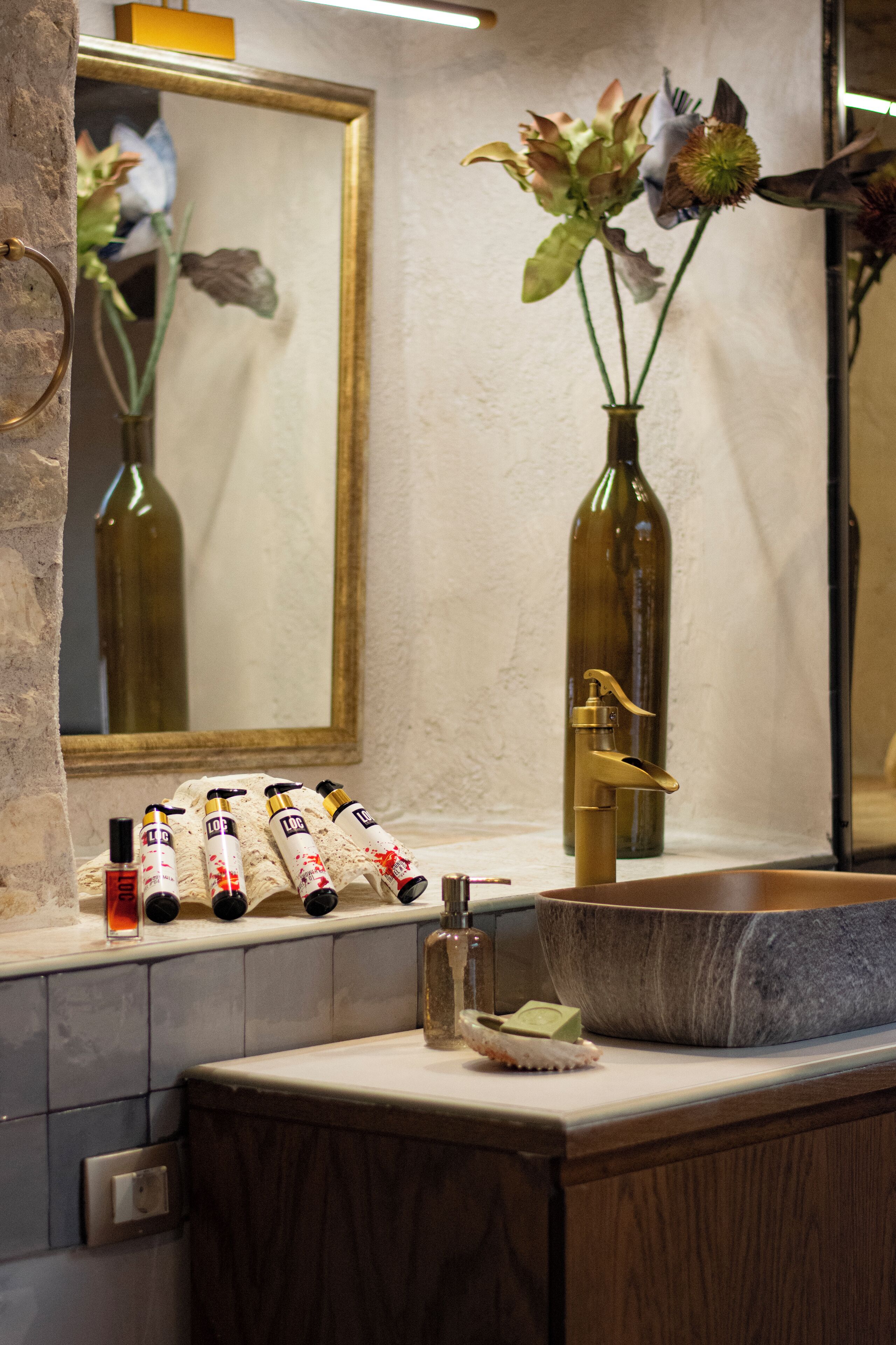A bathroom with a marble sink, a mirror, and a vase with flowers. There are toiletries on the counter.