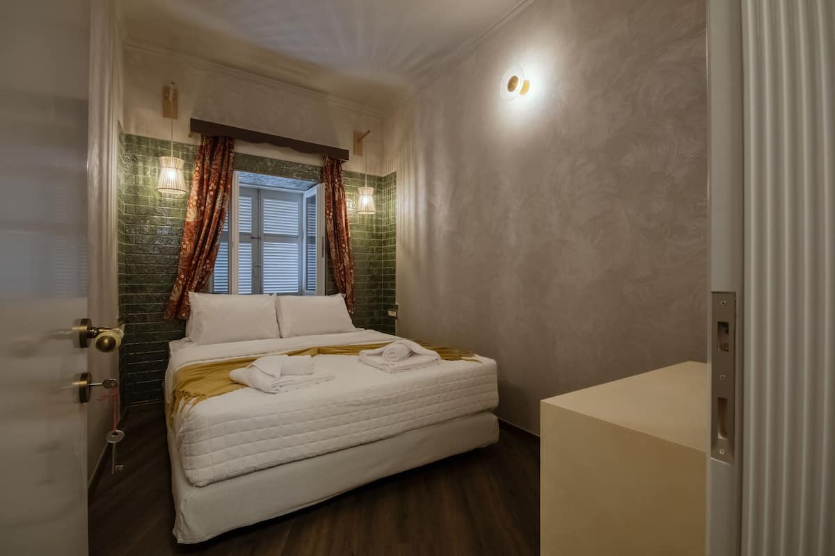 Ein Hotelzimmer mit einem Bett, einem Fenster mit Vorhängen, einer Wandleuchte und einer Tür mit Schlüssel im Schloss.