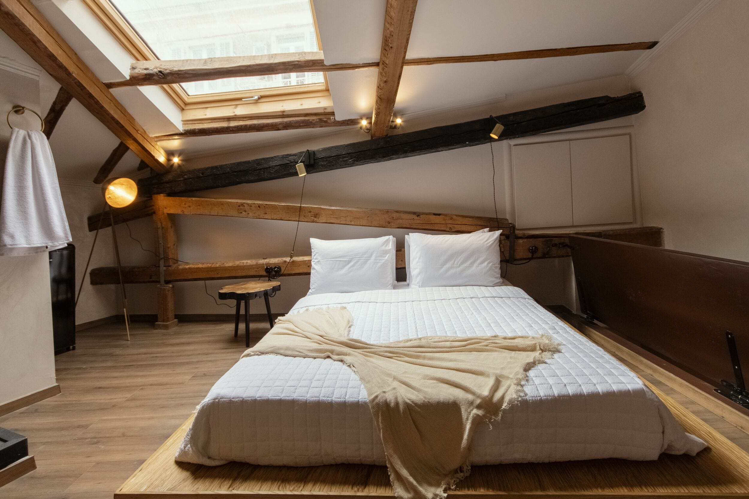 A bedroom with a skylight, wooden beams, a bed with white bedding, and a small table.