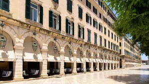 Una fila de asientos al aire libre con columnas y arcos de piedra, un camino empedrado y un edificio con varias ventanas y balcones.