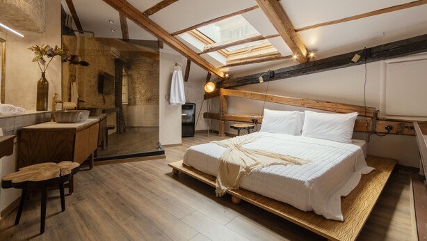 Un dormitorio con techo estilo loft, vigas de madera, una cama con ropa de cama blanca, un escritorio de madera y una pequeña zona de baño.