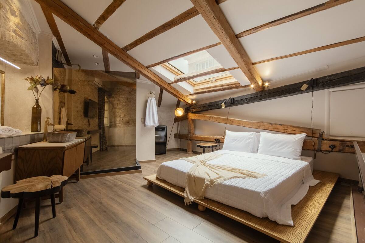 Ein Schlafzimmer mit Dachschrägen, Holzbalken, einem Bett mit weißer Bettwäsche, einem hölzernen Schreibtisch und einer kleinen Badeinrichtung.
