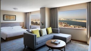 Una camera d'albergo con un letto, un divano grigio, un tavolino da caffè e vista sulla città e sul mare.