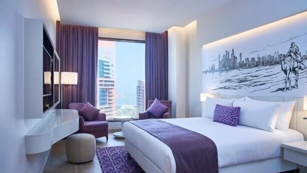 Ein modernes Hotelzimmer mit einem großen Bett, einer Sitzecke mit Stuhl und Hocker, einem Stadtbild-Wandgemälde und Blick auf hohe Gebäude durch das Fenster.