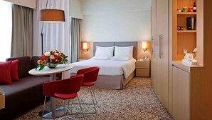 Ein Hotelzimmer mit einem Bett, einem Schreibtisch mit Stühlen, einer Couch, einer Lampe und einem Regal mit Gegenständen.