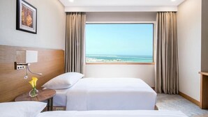 Ein Hotelzimmer mit zwei Betten, einem großen Fenster mit Blick auf den Strand, einem Holz-Kopfteil und einem kleinen Tisch mit einer Blumenvase.