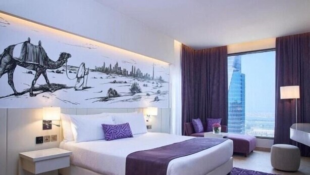A modern hotel room with a large bed, a nightstand, a seating area with a chair and ottoman, a large window with a city view, and a camel illustration on the wall. - Two Bedroom Suite Near Dic Metro (Dubai)