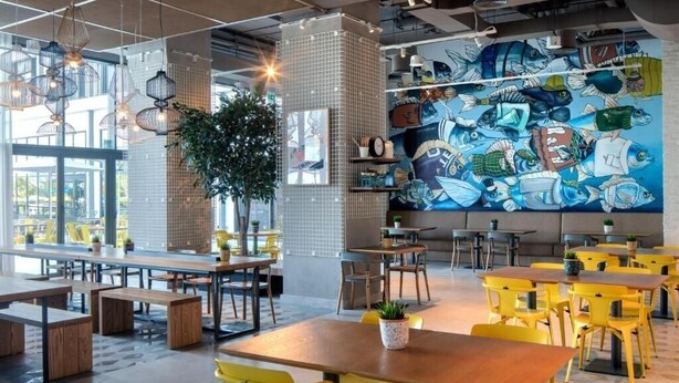 Un intérieur de restaurant moderne avec des chaises jaunes, des tables en bois et une fresque représentant des poissons sur le mur.
