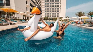 Une piscine avec des chaises longues et des parasols, un grand flotteur en forme de cygne, et un bâtiment d'hôtel en arrière-plan.