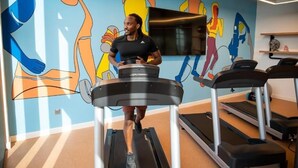 Une personne court sur un tapis de course dans une salle de sport, et un mur coloré orné d’une fresque se trouve à proximité.