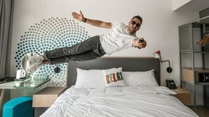 Une personne sautant dans une chambre d'hôtel moderne, qui contient un lit, une table de chevet, un bureau et un mur orné d'un motif à pois.