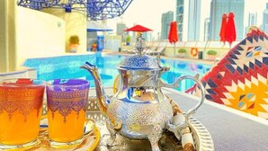 A table with a teapot and glasses, a blue geometric umbrella, and a cityscape with tall buildings in the background. - Deluxe Room Near Burj Khalifa Metro By E R (Dubai)