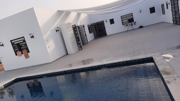 A swimming pool with a curved white wall and windows with metal shutters. - Maison D'hote Saida Djerba Houmt Souk (Sedghiane)
