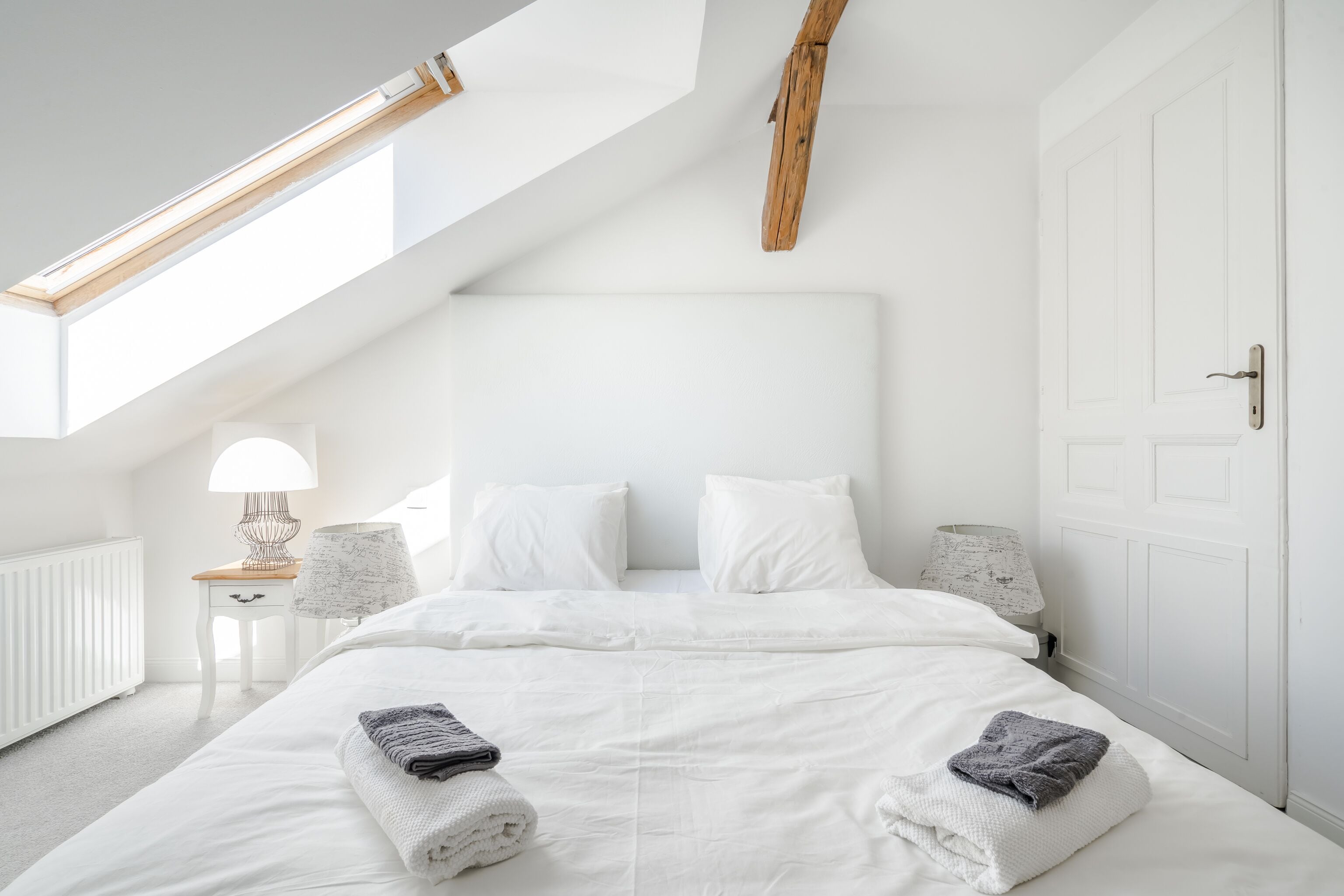 A bedroom with a skylight, white bedding, a wooden headboard, and a bedside lamp.