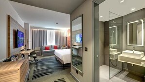 A modern hotel room with a large bed, a desk, a TV, a sofa, and a separate bathroom with a glass shower. - Standard Room Near Sharjah Expo Centre By E R (Sharjah)