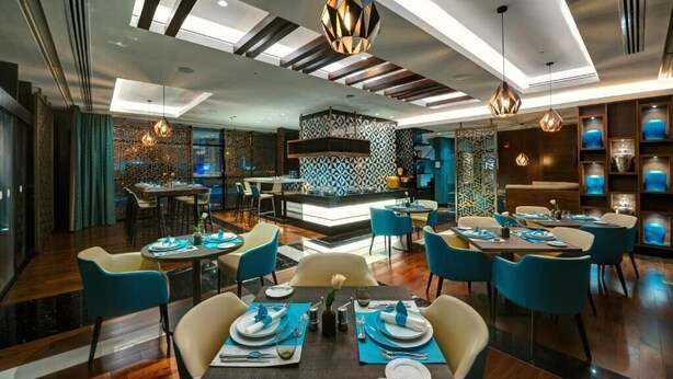 A modern dining area with blue chairs, wooden tables, and geometric patterned walls. - Standard Room Near Sharjah Expo Centre By E R (Sharjah)