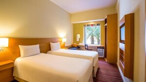 Habitación de hotel con dos camas, un escritorio, una silla y una ventana con vistas a árboles.