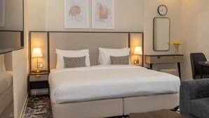 Une chambre d’hôtel avec un grand lit, deux tables de chevet avec des lampes, un bureau avec une chaise, un miroir et deux œuvres d’art encadrées accrochées au mur.