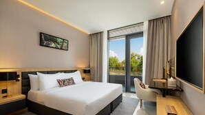 Ett modernt hotellrum med en stor säng, ett skrivbord med en platt-TV, en stol och en balkong med utsikt över grönområden.