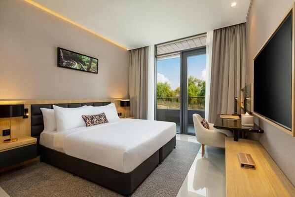 Ein modernes Hotelzimmer mit einem großen Bett, einem Schreibtisch, einem Flachbildfernseher, einem Sessel und einem Balkon mit Blick auf Grünflächen.