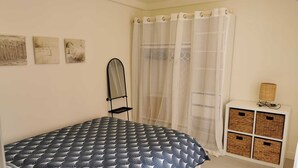 Un dormitorio con una cama, una silla, un mueble bajo con cestas y una cortina.