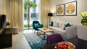 A modern hotel room with a sofa, armchair, coffee table, and a dining table with a bowl of fruit. There is a TV, a lamp, and artwork on the wall. - One Bedroom Apartment Near Moe By E R (Dubai)