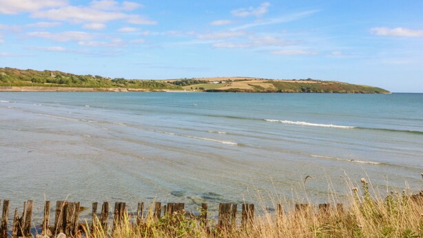 Un paysage côtier avec une plage de sable, une clôture en bois et une colline lointaine couverte d’une végétation clairsemée.