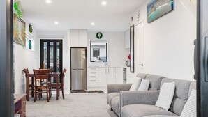 A modern living room with a grey sofa, wooden dining table, and chairs. There is a kitchen area with white cabinets and a stainless steel refrigerator. - Stunning 2BR in Kelston with WIFI and Carpark (Auckland West)