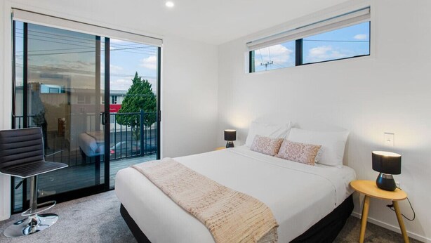 A modern bedroom with a large bed, a glass door leading to a balcony, and a small table with a lamp. - Stunning 2BR in Kelston with WIFI and Carpark (Auckland West)