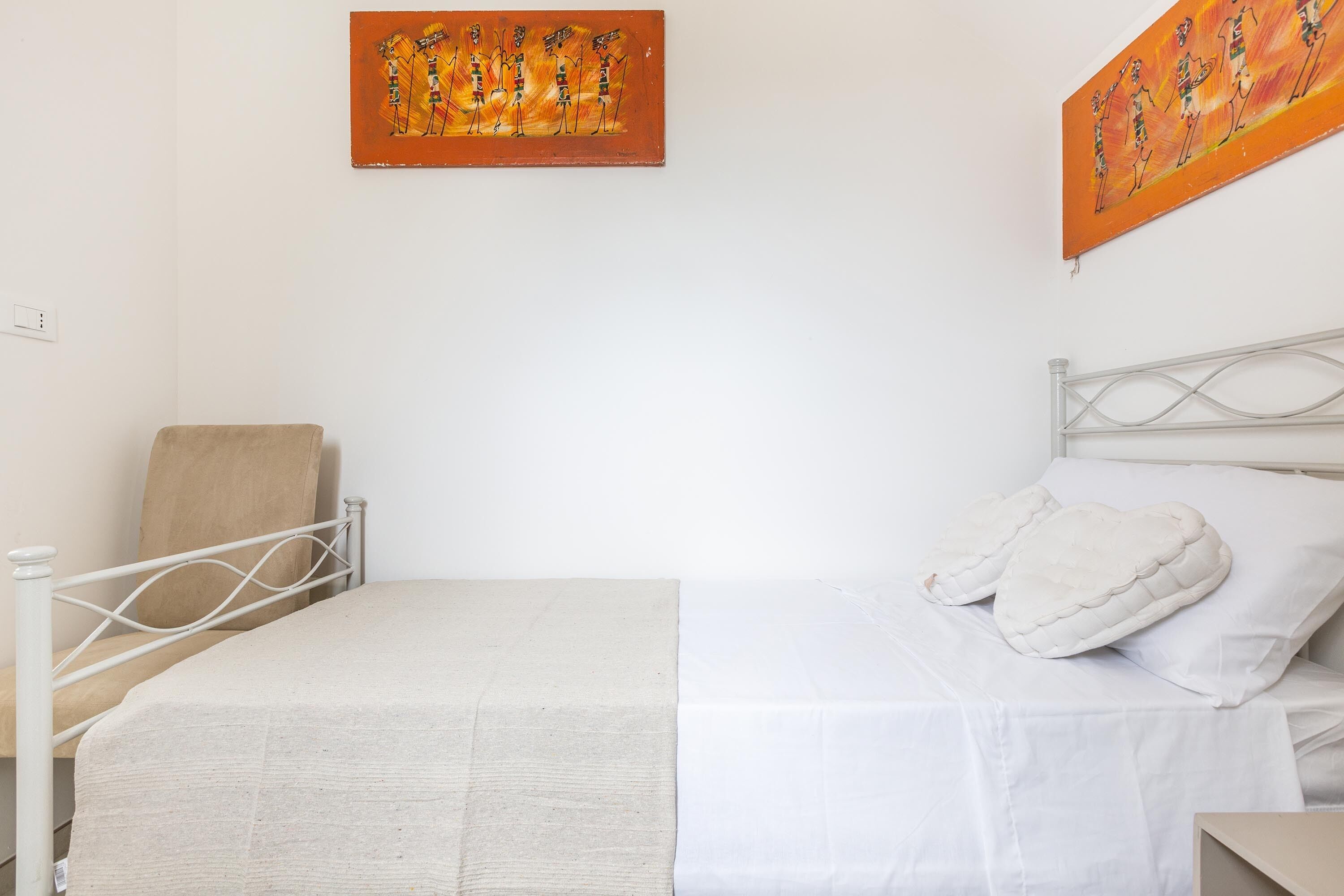 A single bed with a white mattress and a white headboard. There is a beige chair against the wall and a painting above the bed.