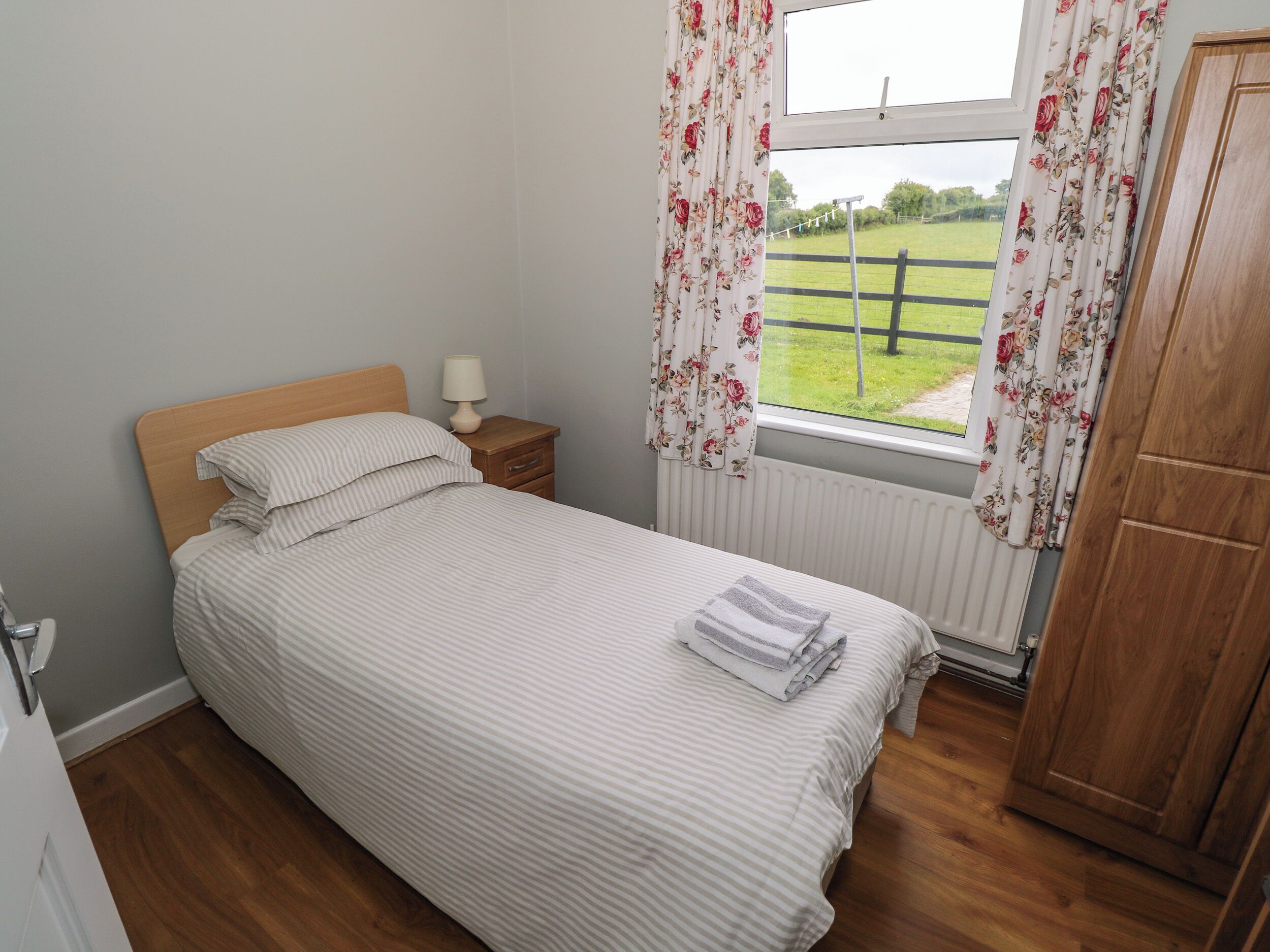 A neatly made bed with striped bedding, a bedside table with a lamp, a window with floral curtains, and a wooden wardrobe.