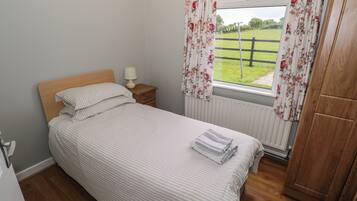 A neatly made bed with striped bedding, a bedside table with a lamp, a window with floral curtains, and a wooden wardrobe.