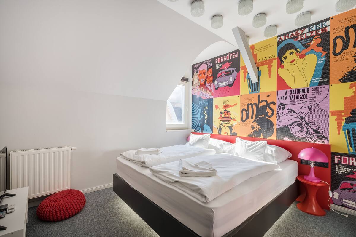 A bedroom with a bed, a red ottoman, a radiator, and a wall with colorful posters.
