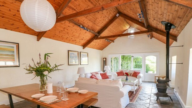A spacious room with a high wooden ceiling, a dining table set with plates and glasses, a white sofa with red pillows, and a stone-tiled floor.