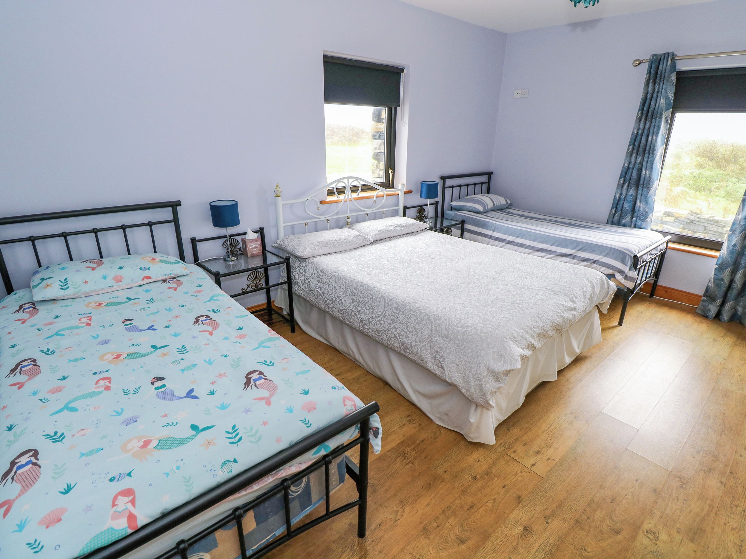 A bedroom with two single beds, a window with curtains, a bedside table with a lamp, and a wall-mounted shelf.