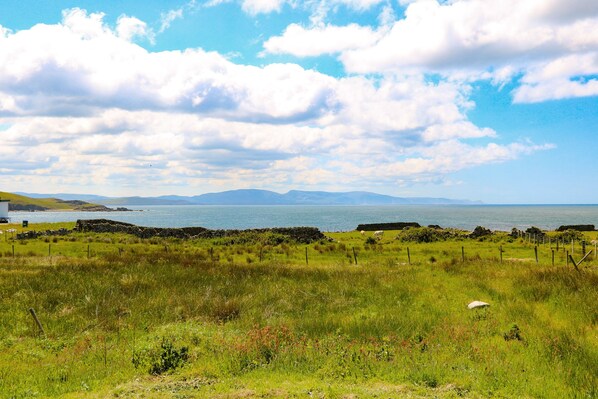 A coastal landscape with a grassy field, a stone wall, and a view of the sea and distant mountains.