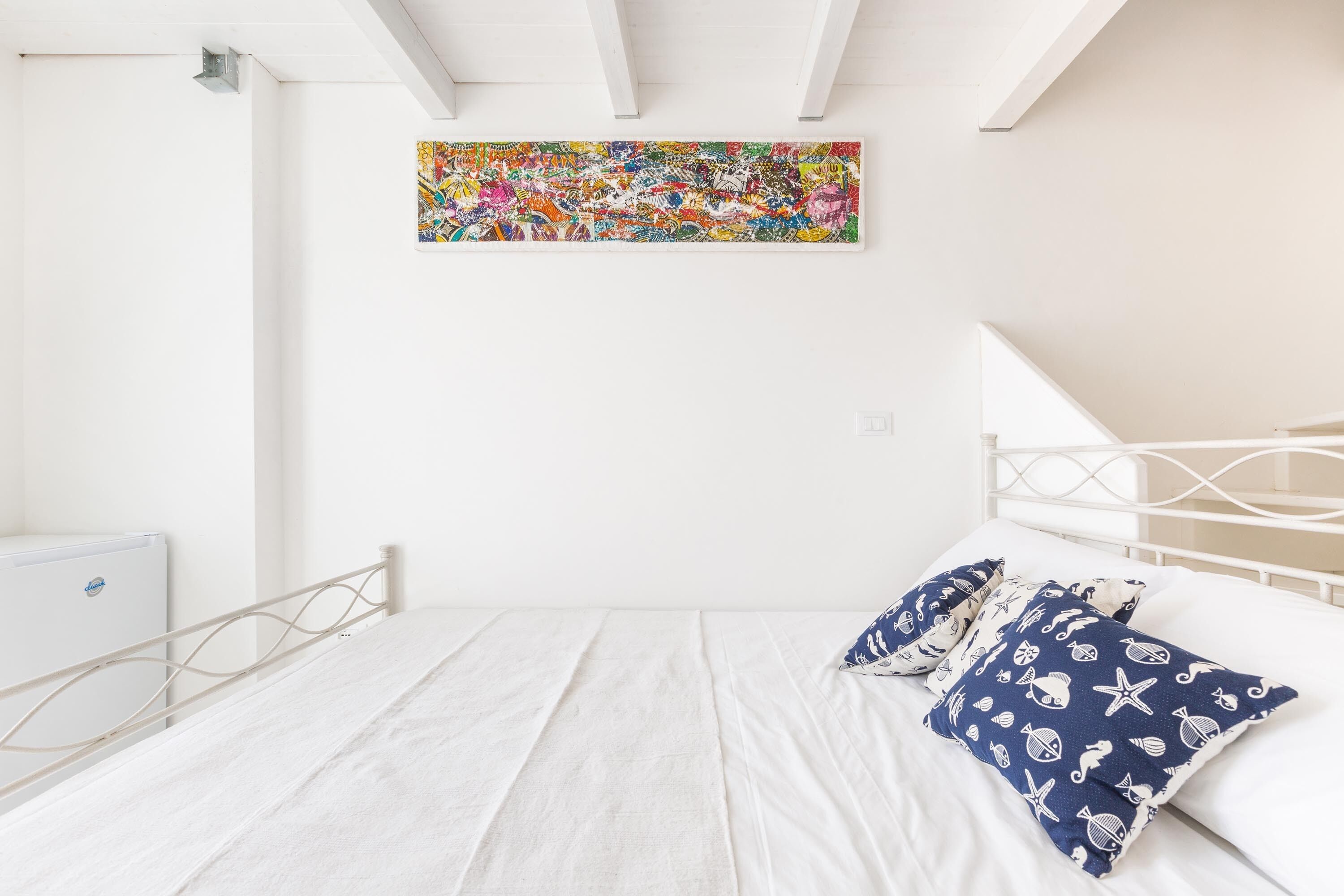Ein Schlafzimmer mit einem weißen Bett, einem weißen Kopfteil und einem farbenfrohen, abstrakten Gemälde an der Wand.