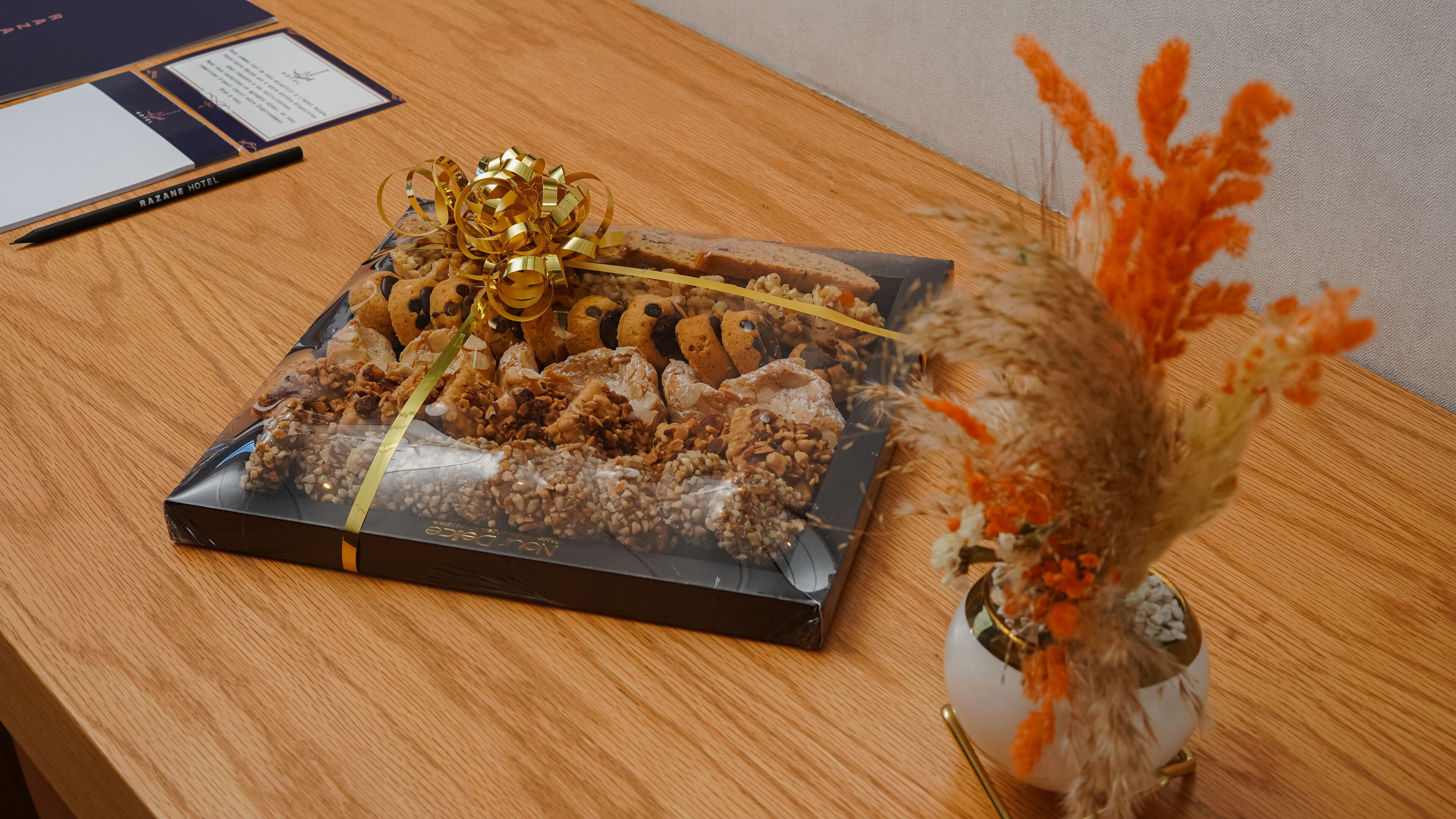 Una mesa de madera con una caja de regalo que contiene galletas y un lazo dorado, y un jarrón con flores naranjas.