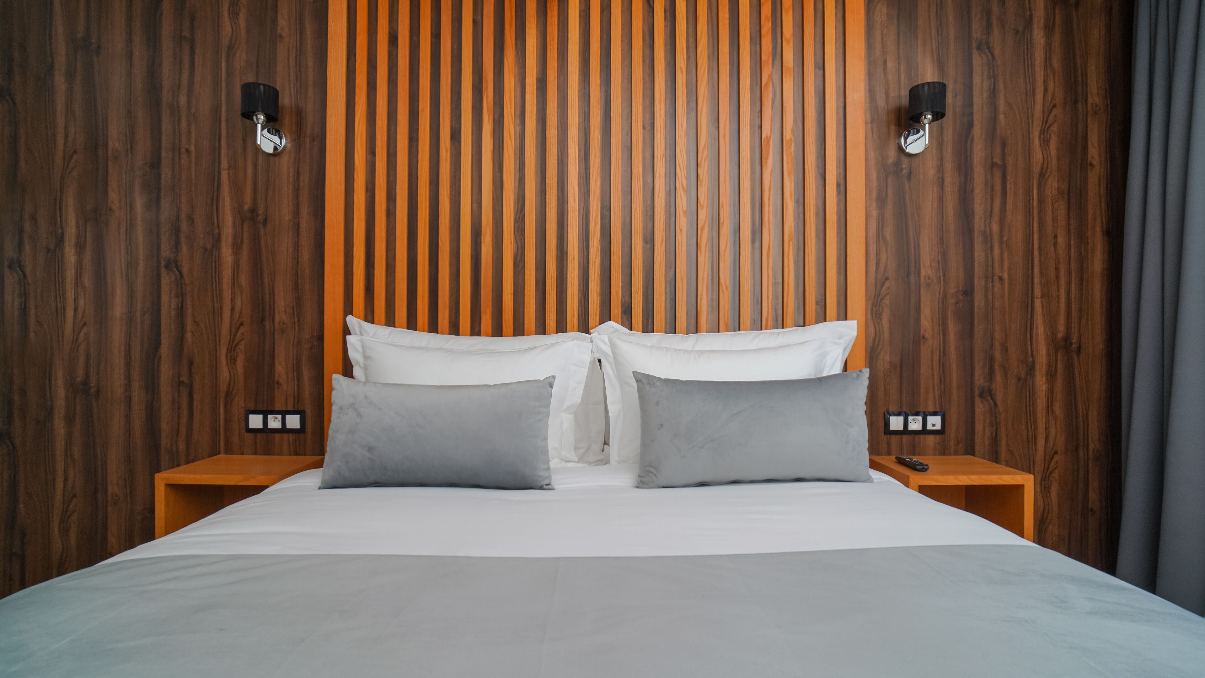 Una cama bien hecha con ropa de cama blanca y gris, cabecera de madera con listones verticales, dos mesitas de noche con lámparas y un enchufe en la pared.