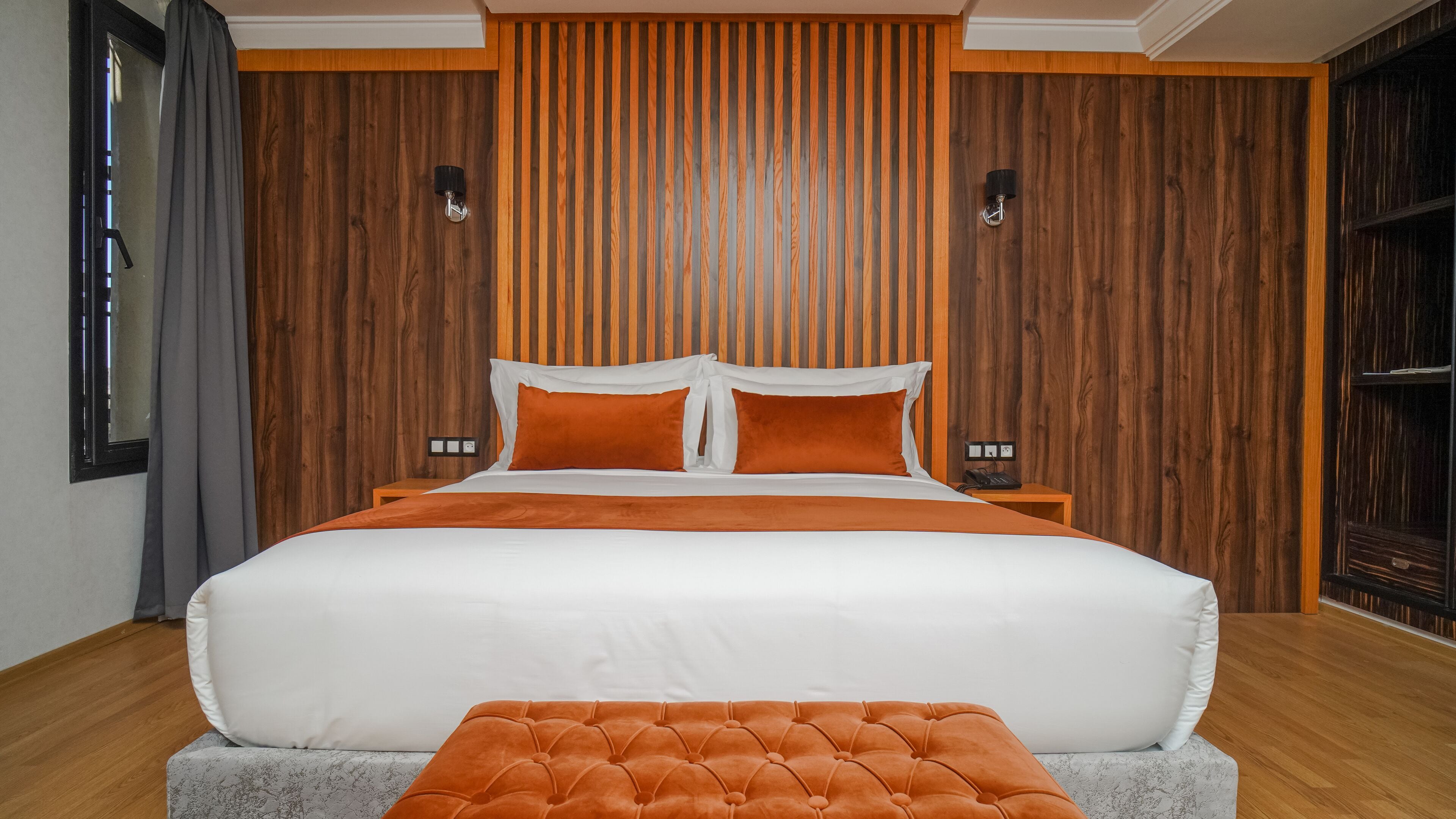 A large bed with white and orange bedding, a tufted orange ottoman, and wooden panel walls.