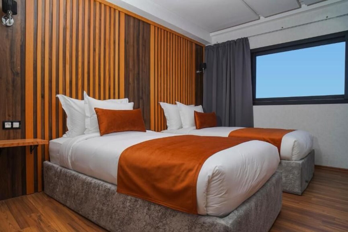 Cama doble con ropa de cama blanca y naranja, pared de acento de madera y ventana con cortinas.