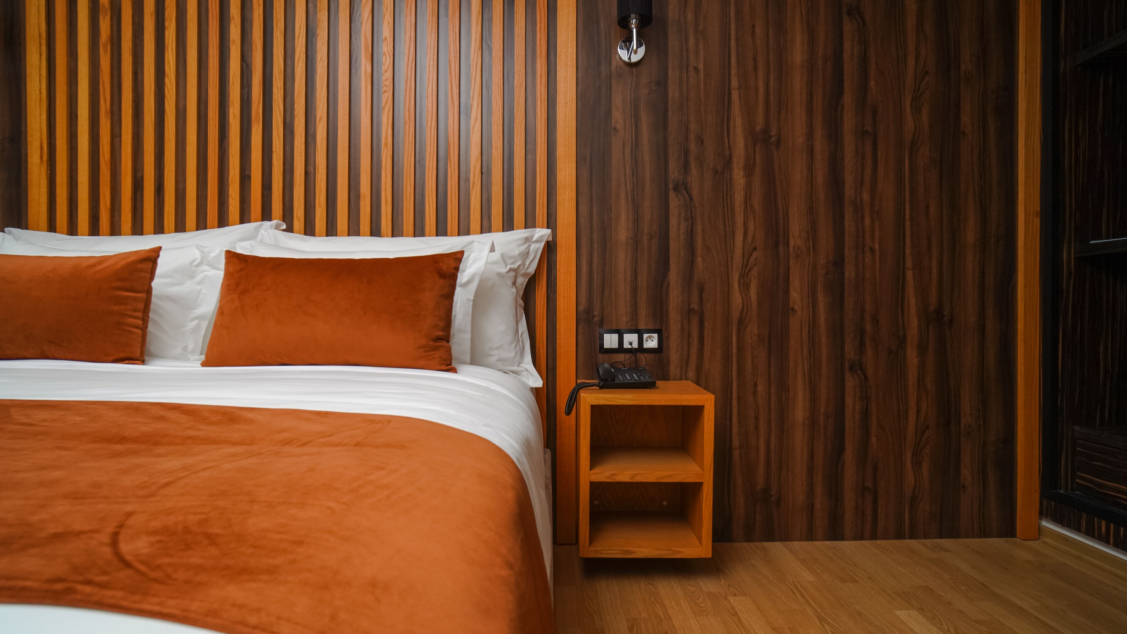 A neatly made bed with white and orange bedding, a wooden nightstand with a phone, and a wall with vertical wooden slats.