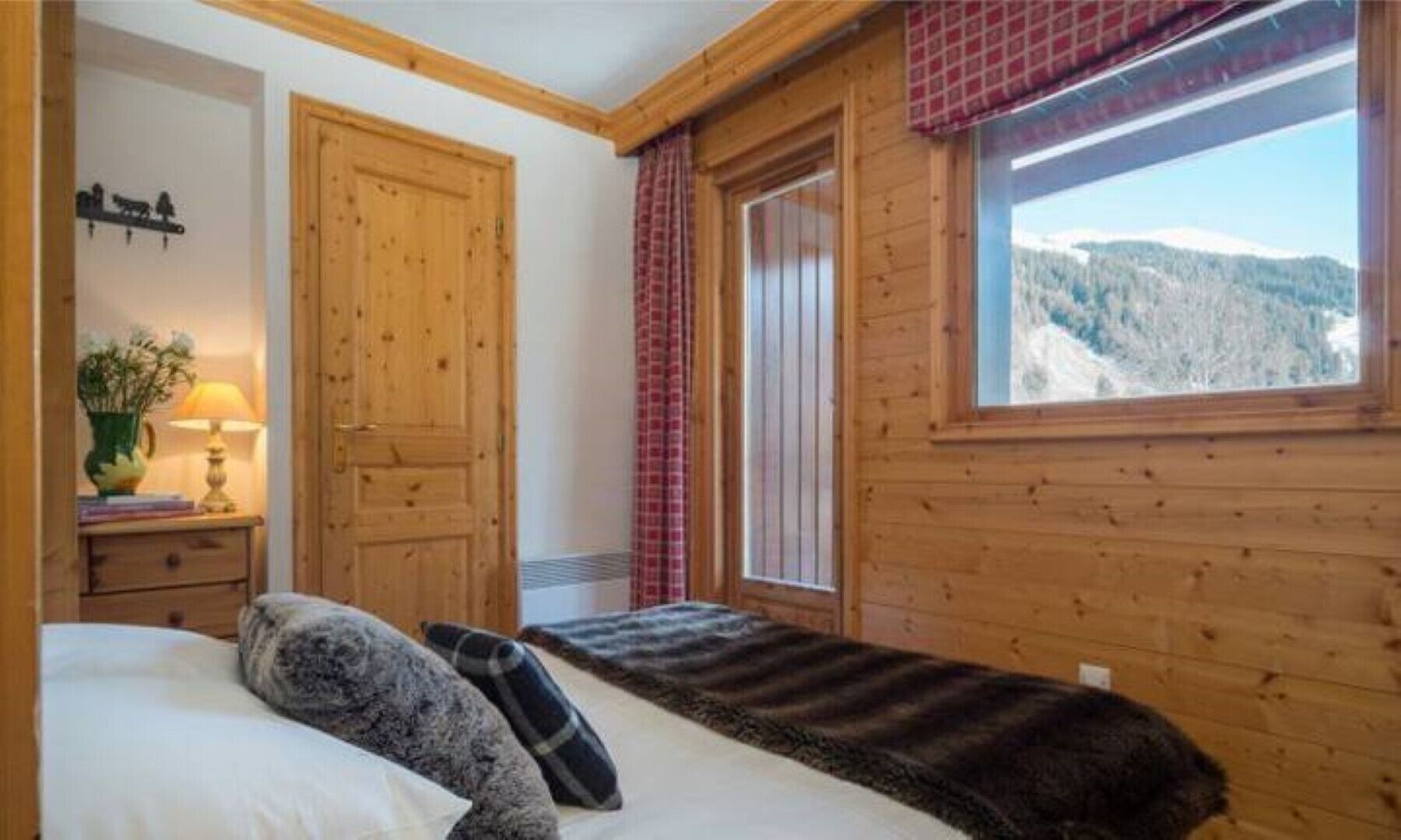 A wooden cabin room with a bed, a nightstand with a lamp, a window with a view of snow-covered mountains, and a door.