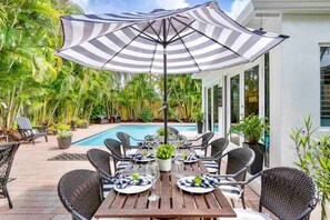 An outdoor dining area with a pool, surrounded by lush greenery and a large striped umbrella.