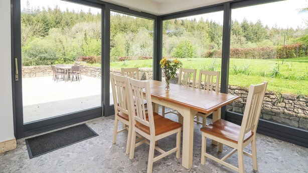 A dining area with a wooden table and chairs, large windows offering a view of a garden.