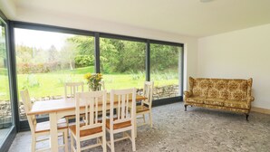 A room with a dining table and chairs, a sofa, and large windows offering a view of a garden.