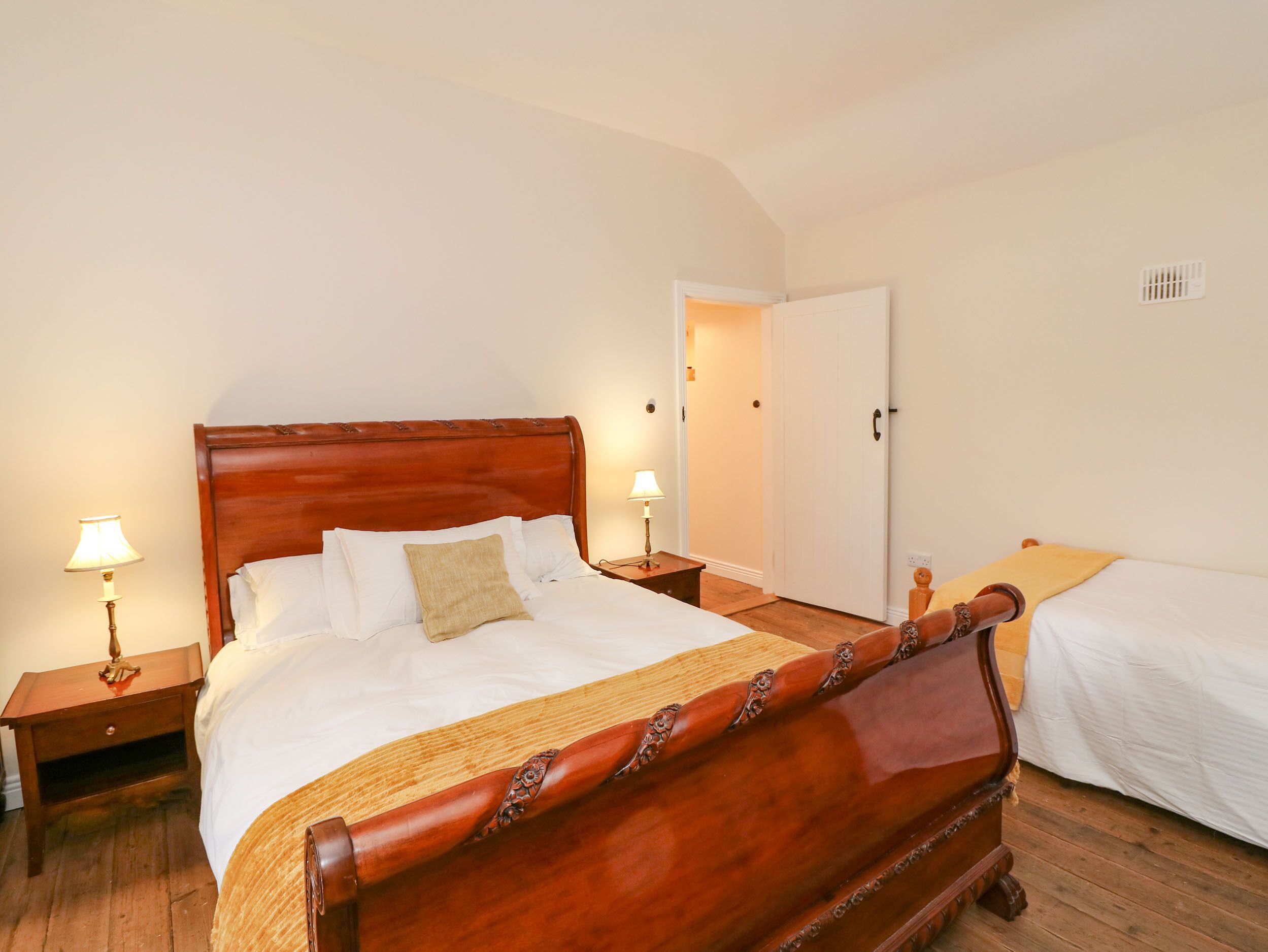 A wooden four-poster bed with white linens and a beige throw pillow. There are two bedside tables with lamps, a wooden floor, and a white door in the background.